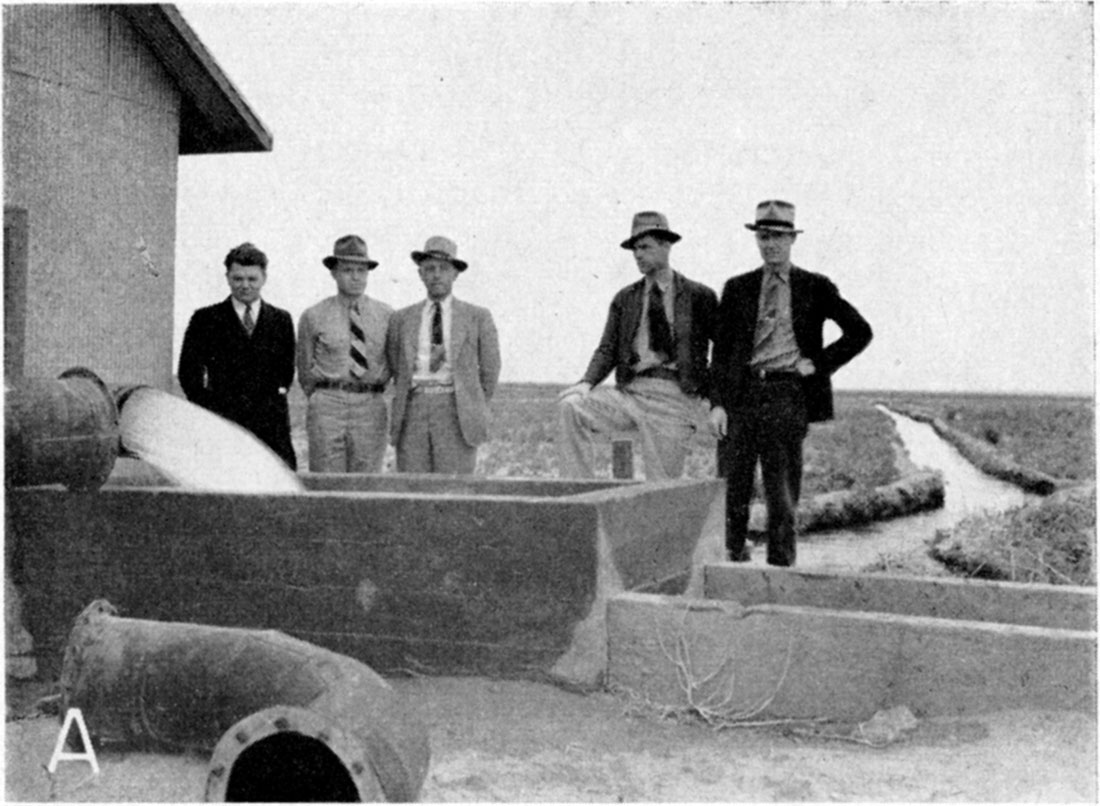 Black and white photo: five men next to horizontal pipe with water going into cement tank,