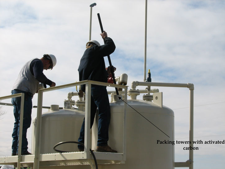 Two men on platform attached to tanks are tamping down carbon with a pole.